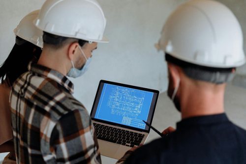 Civil engineers wearing hard hats reviewing building blueprints on a laptop at a construction site, collaborating on project plans.