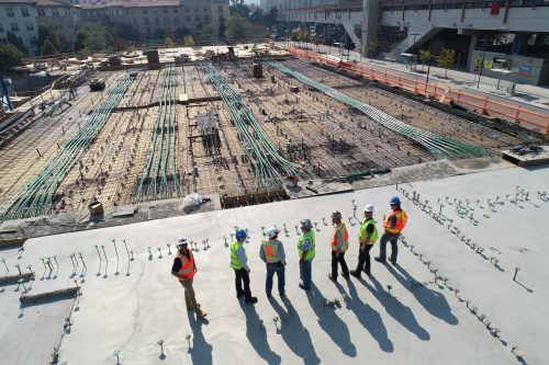 Group of engineers and project planners standing on the roof of a half-finished construction site. They are overlooking a large-scale project with exposed concrete and metal rebar protruding from the ground, preparing for further concrete work.